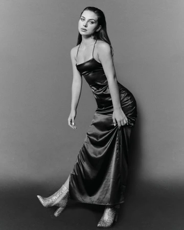 Monochrome studio portrait of a young woman in a satin dress and glitter boots, posing and looking at the camera with a neutral expression.