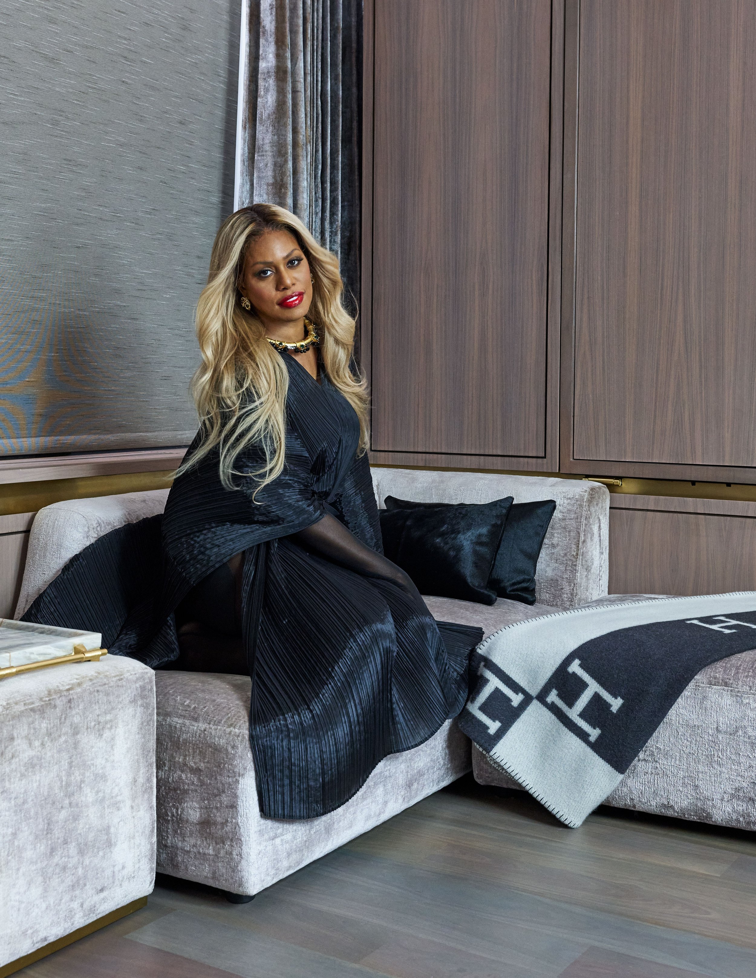 Woman in a black, shimmering dress poses on a taupe chaise lounge with a black and gray blanket, set against a textured wall.