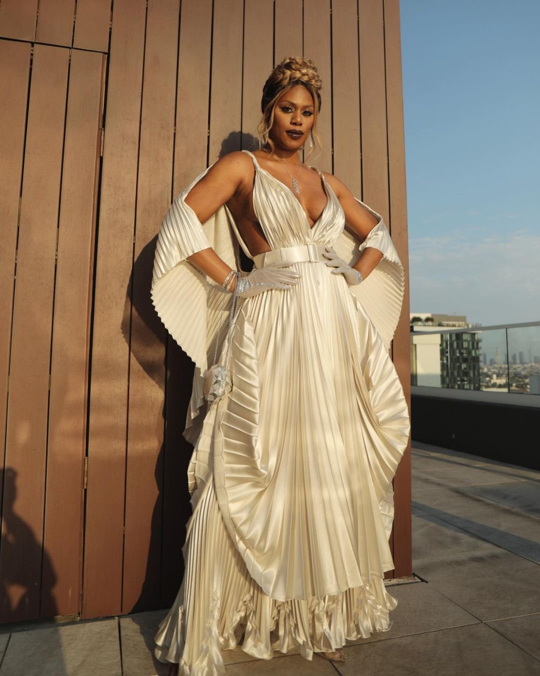 Elegant woman in pleated gown with opera gloves poses on a rooftop with city buildings in the background in an eye-level shot.