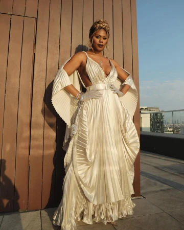 Elegant woman in pleated gown with opera gloves poses on a rooftop with city buildings in the background in an eye-level shot.