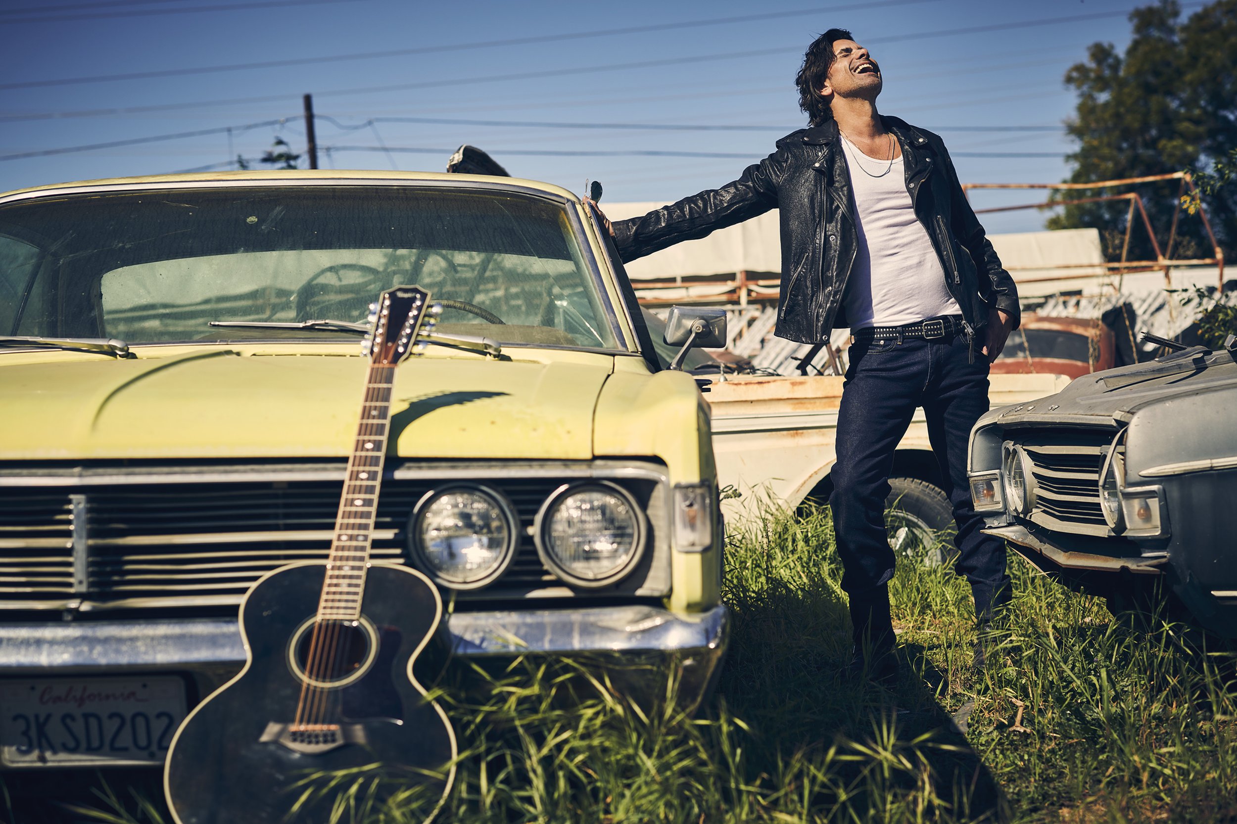 Adult male in a black leather jacket leans against a yellow vintage car with a guitar, set in an outdoor junkyard-like environment.