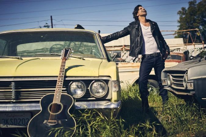 Adult male in a black leather jacket leans against a yellow vintage car with a guitar, set in an outdoor junkyard-like environment.