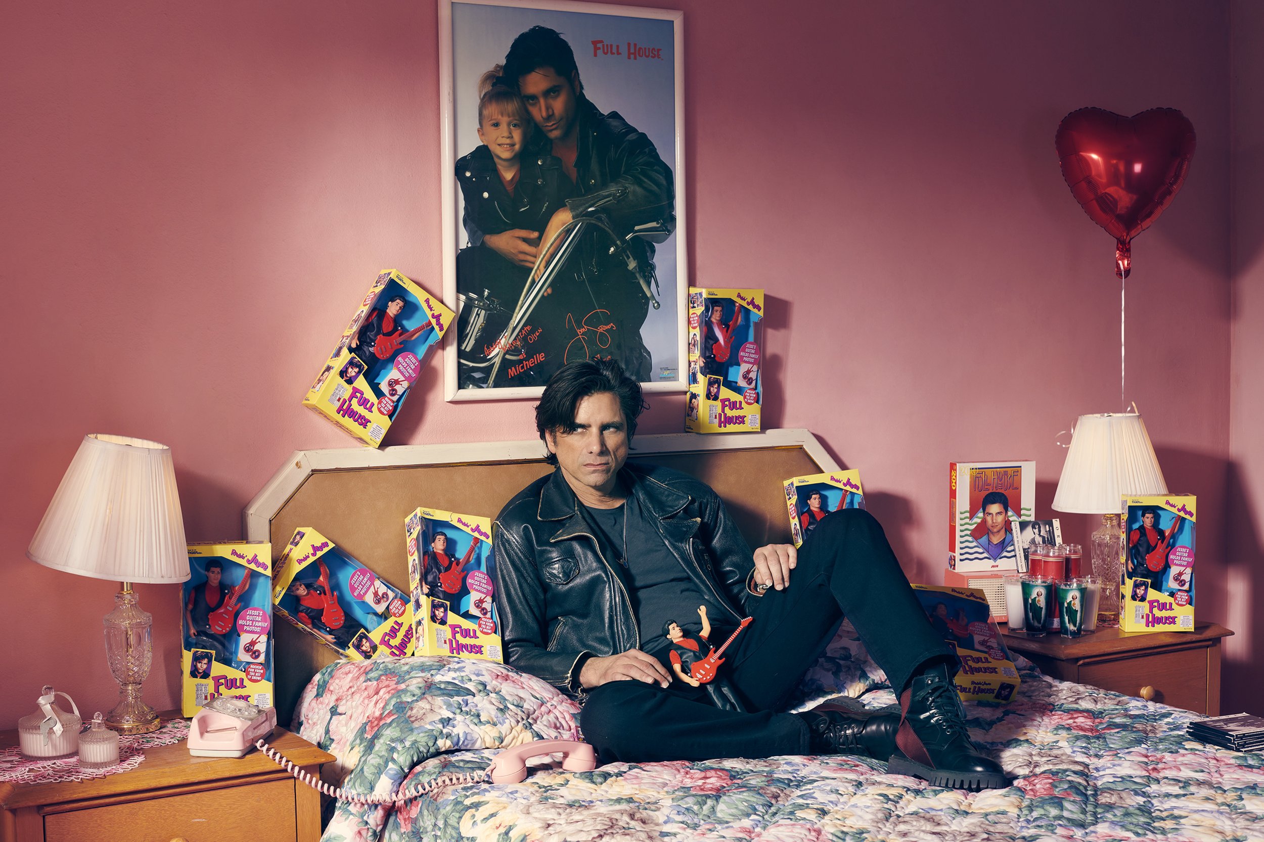 Adult male poses on a bed in a pink room surrounded by Full House dolls and memorabilia, including a poster and other objects.