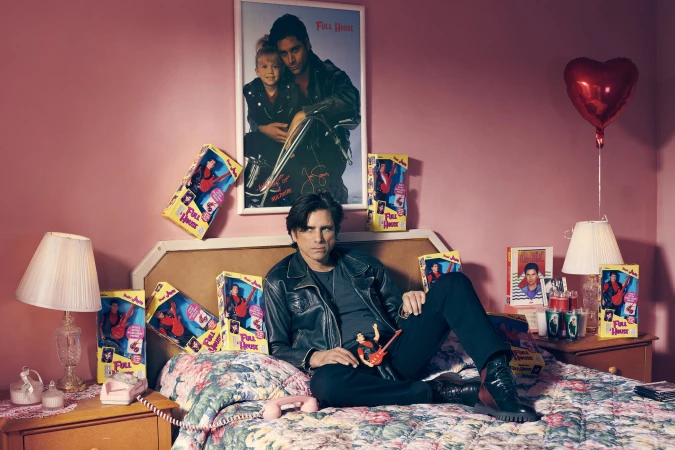 Adult male poses on a bed in a pink room surrounded by Full House dolls and memorabilia, including a poster and other objects.
