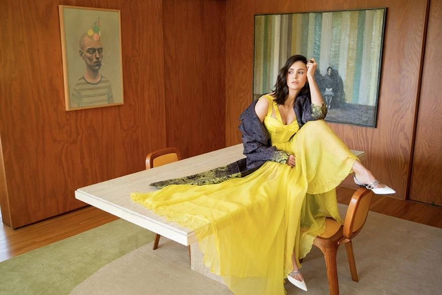 A woman in a yellow dress sits on a table in a room with wood paneling, artwork, and a light green rug, bathed in natural light.