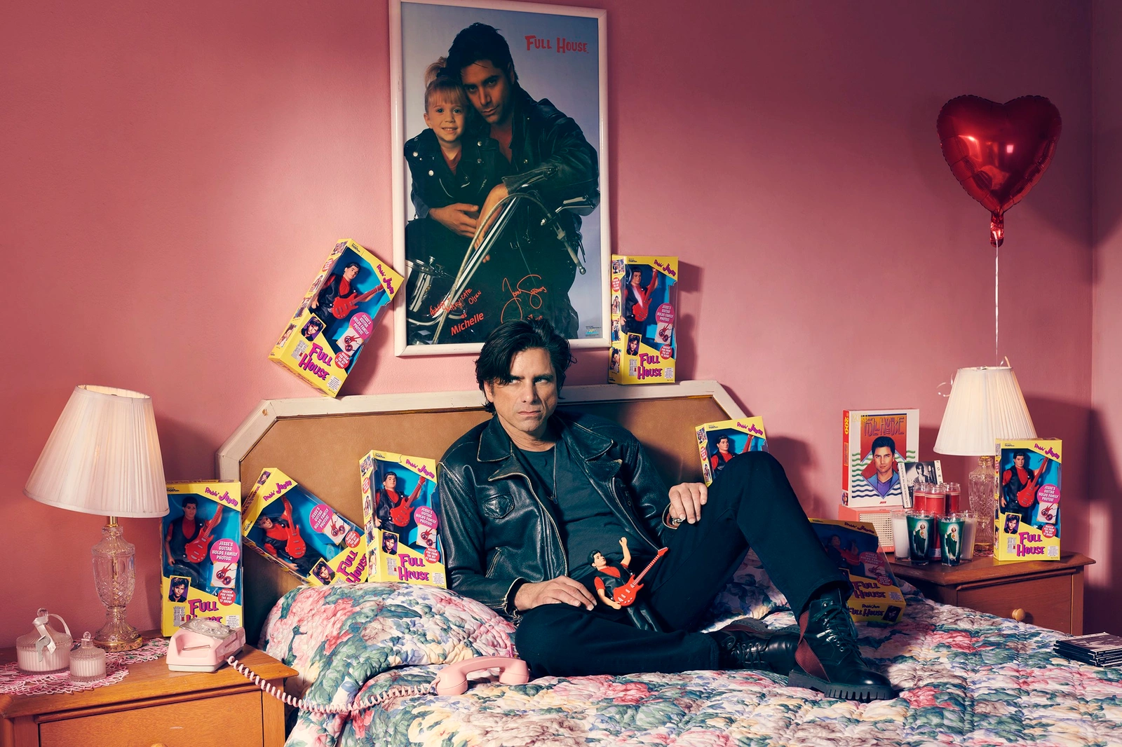 Man wearing a black leather jacket seated on a floral bed surrounded by Full House memorabilia and a large framed poster