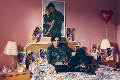 Man wearing a black leather jacket seated on a floral bed surrounded by Full House memorabilia and a large framed poster