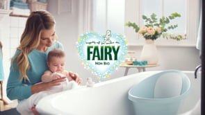 Blond woman in a blue shirt holds a baby near a white bathtub with a baby bathtub insert, flowers, and a Fairy logo. Indoor shot.
