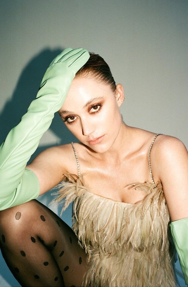 Model wearing a feather-trimmed dress, polka dot tights, and long green leather gloves posing against a grey backdrop