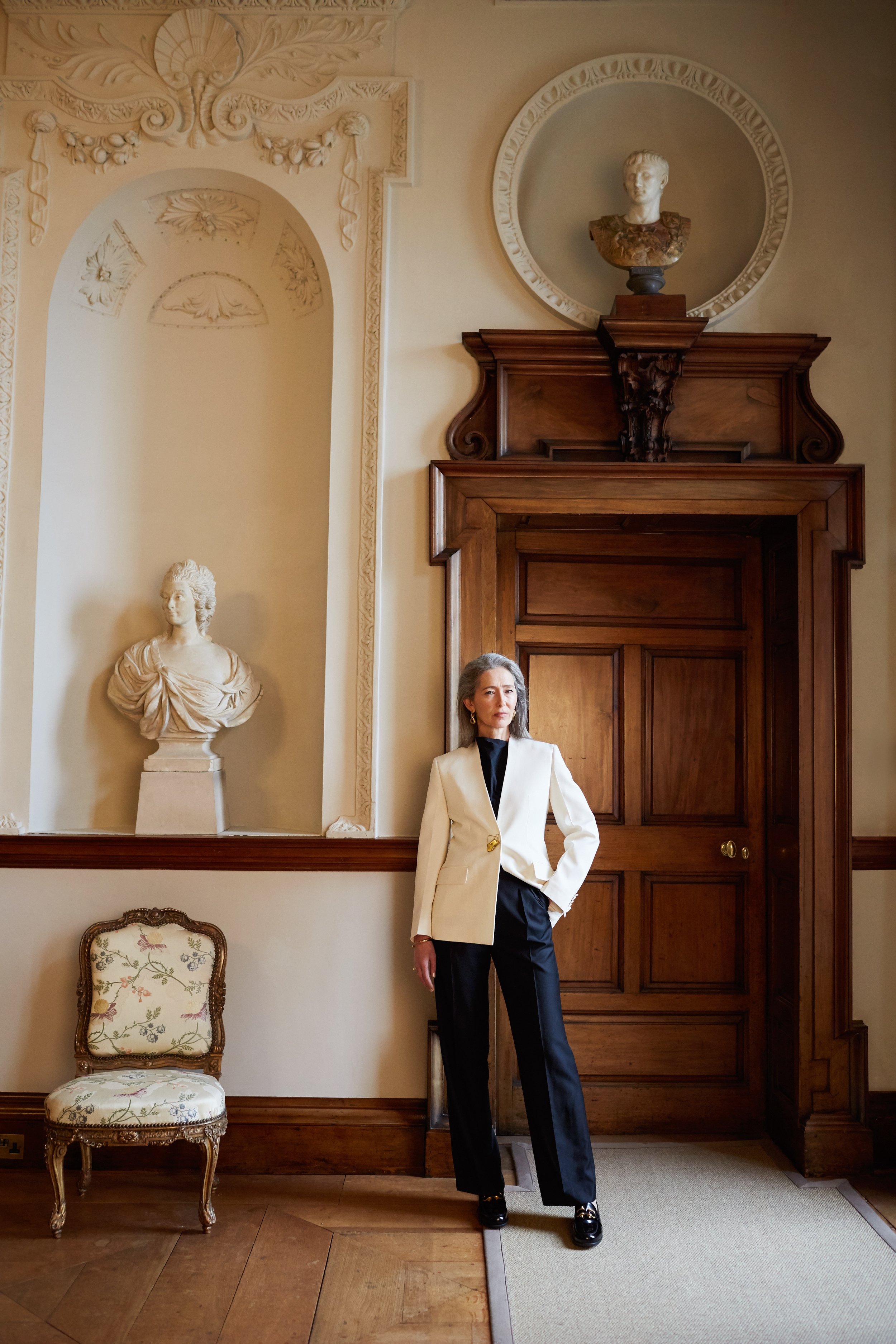 Elegant woman in a cream suit standing in an ornate hallway with classical statues, detailed woodwork, and polished wood floors.