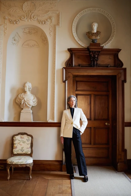 Elegant woman in a cream suit standing in an ornate hallway with classical statues, detailed woodwork, and polished wood floors.