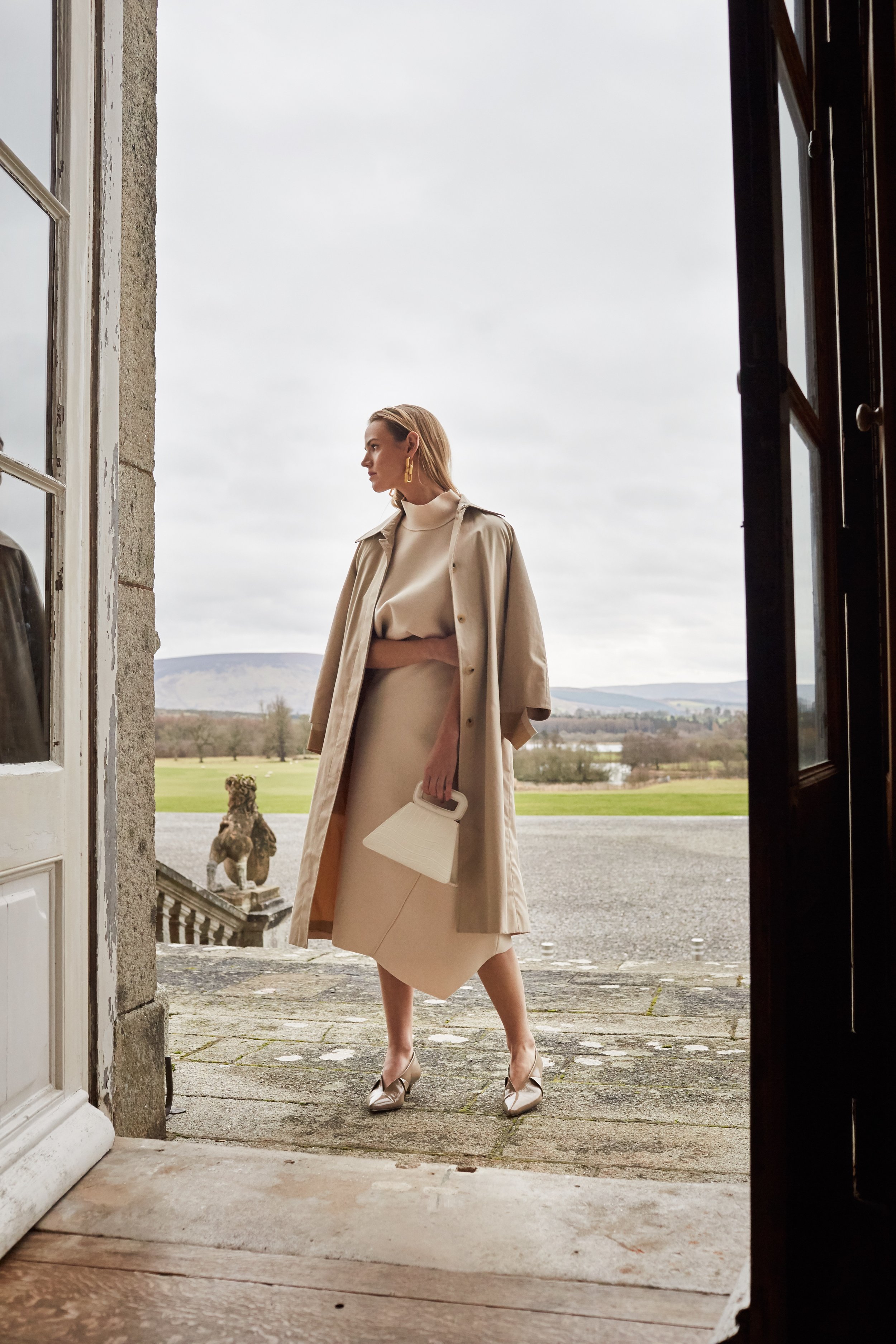 Woman in a beige trench coat and dress stands in a doorway overlooking a green lawn and hills on a cloudy day, holding a purse.