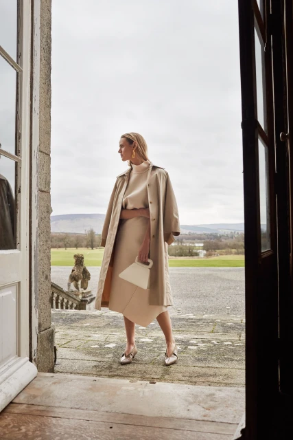 Woman in a beige trench coat and dress stands in a doorway overlooking a green lawn and hills on a cloudy day, holding a purse.