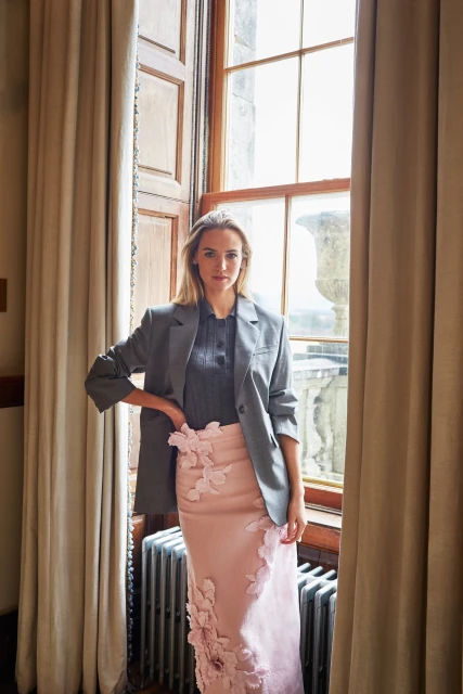 A woman in a blazer and skirt stands near a window, featuring natural light, neutral colors, and drapes, with an indoor setting.