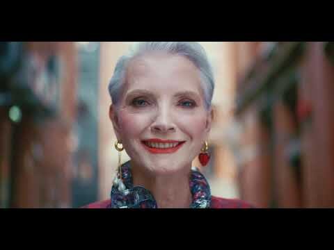 Elegant elderly woman with short gray hair, red lipstick, and heart earrings smiles in a close-up shot on an urban street.