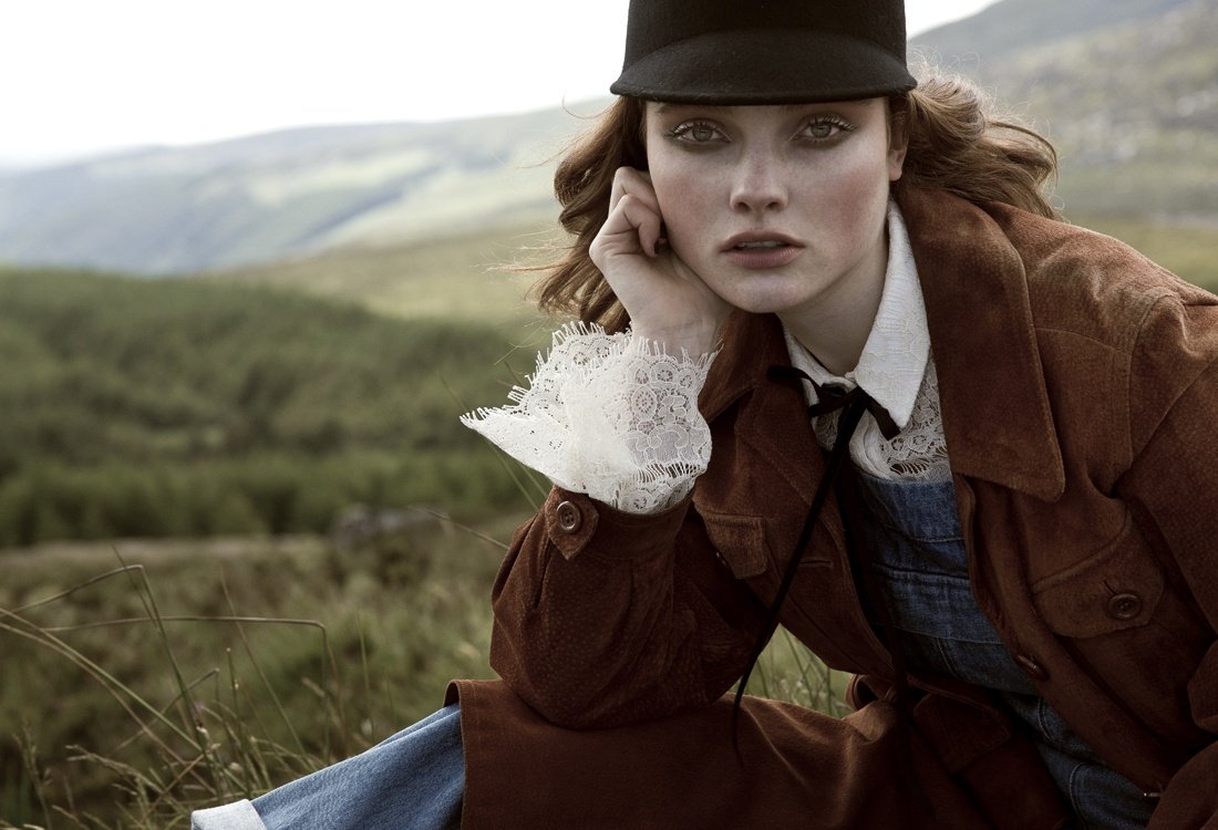 A young woman in a brown jacket and black cap poses in a grassy field with mountains in the background, creating a vintage fashion editorial.