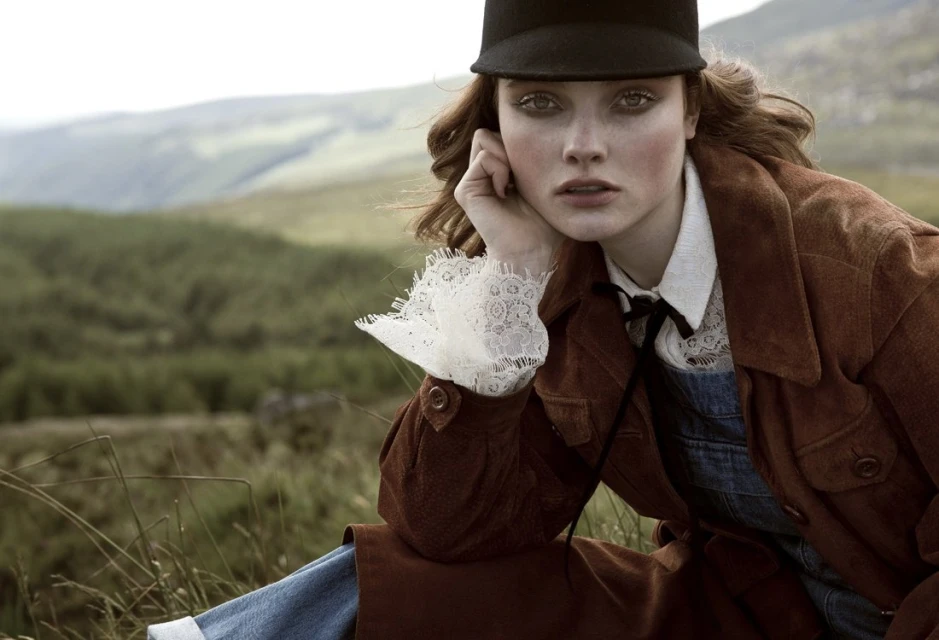 A young woman in a brown jacket and black cap poses in a grassy field with mountains in the background, creating a vintage fashion editorial.