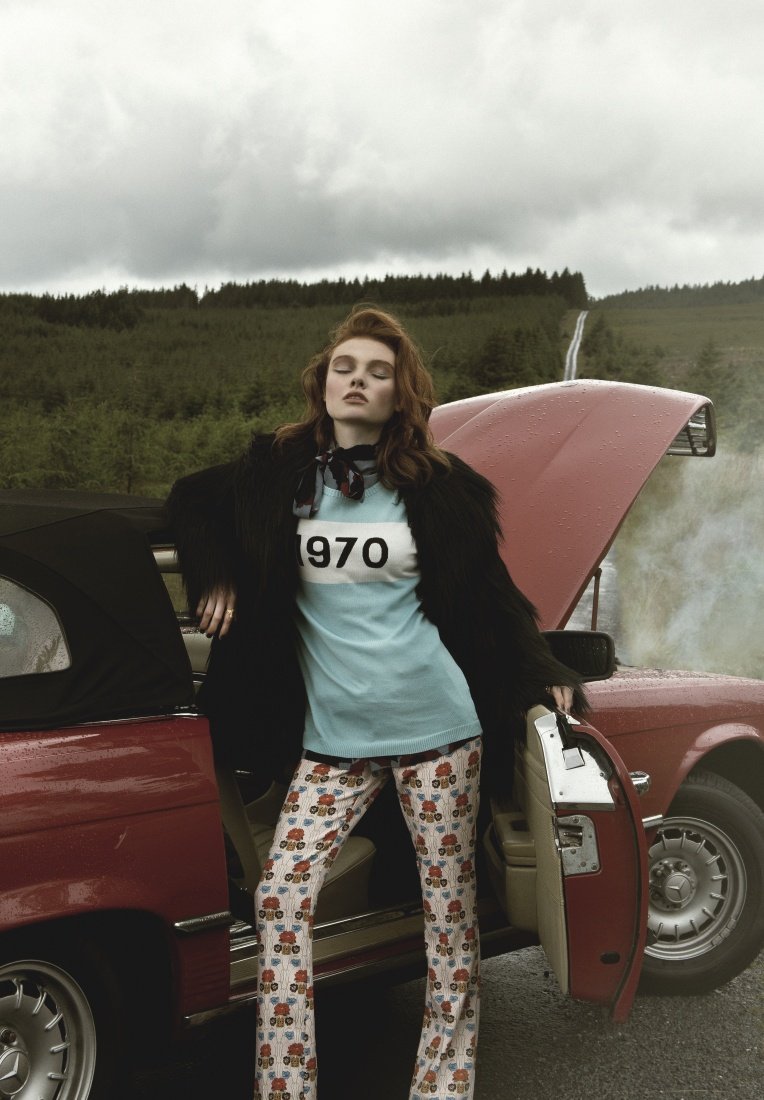 A woman wearing patterned pants and a t-shirt leans against a red vintage car in an outdoor setting, with a forest in the background.