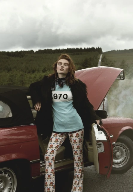 A woman wearing patterned pants and a t-shirt leans against a red vintage car in an outdoor setting, with a forest in the background.