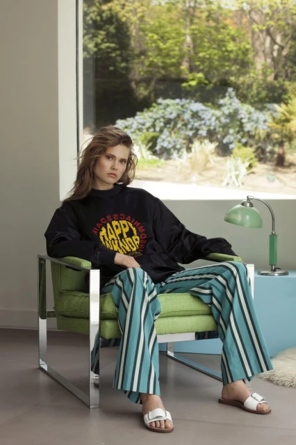 Indoor fashion shot of a woman in a black sweatshirt and striped pants, sitting in a green chair with a window overlooking trees.