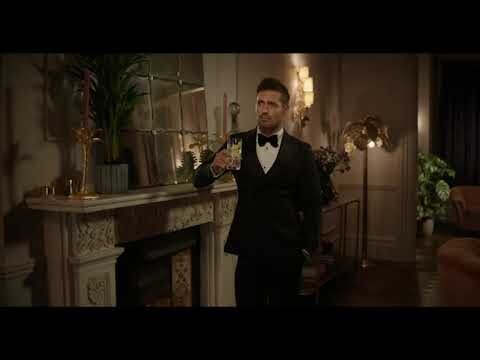 Man in black tuxedo holding glass indoors near fireplace and plants, posing elegantly in a well-lit interior.