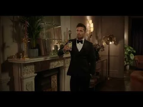 Man in black tuxedo holding glass indoors near fireplace and plants, posing elegantly in a well-lit interior. 