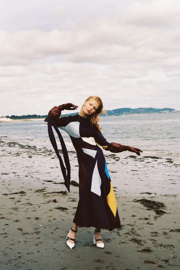 A slender woman in a color-blocked dress and gloves poses dynamically on a sandy beach against a backdrop of sea and cloudy skies.