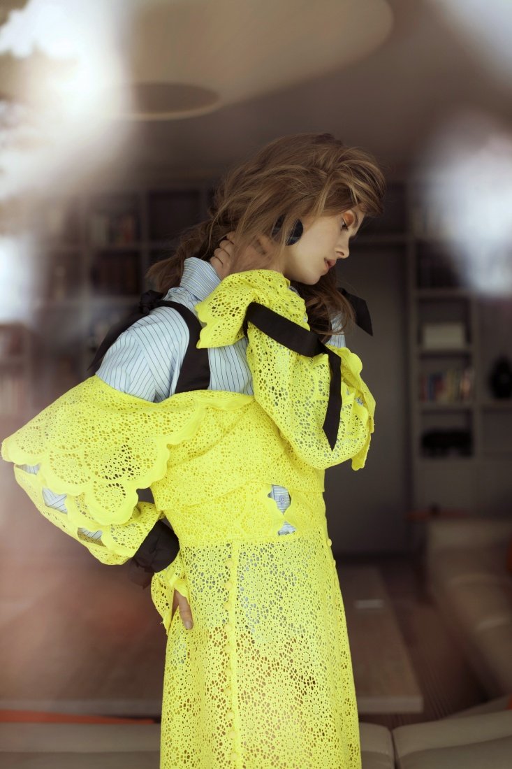 A woman poses indoors wearing a sheer yellow lace dress, layered over a striped shirt with black bows, looking away from the camera.