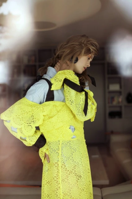 A woman poses indoors wearing a sheer yellow lace dress, layered over a striped shirt with black bows, looking away from the camera.
