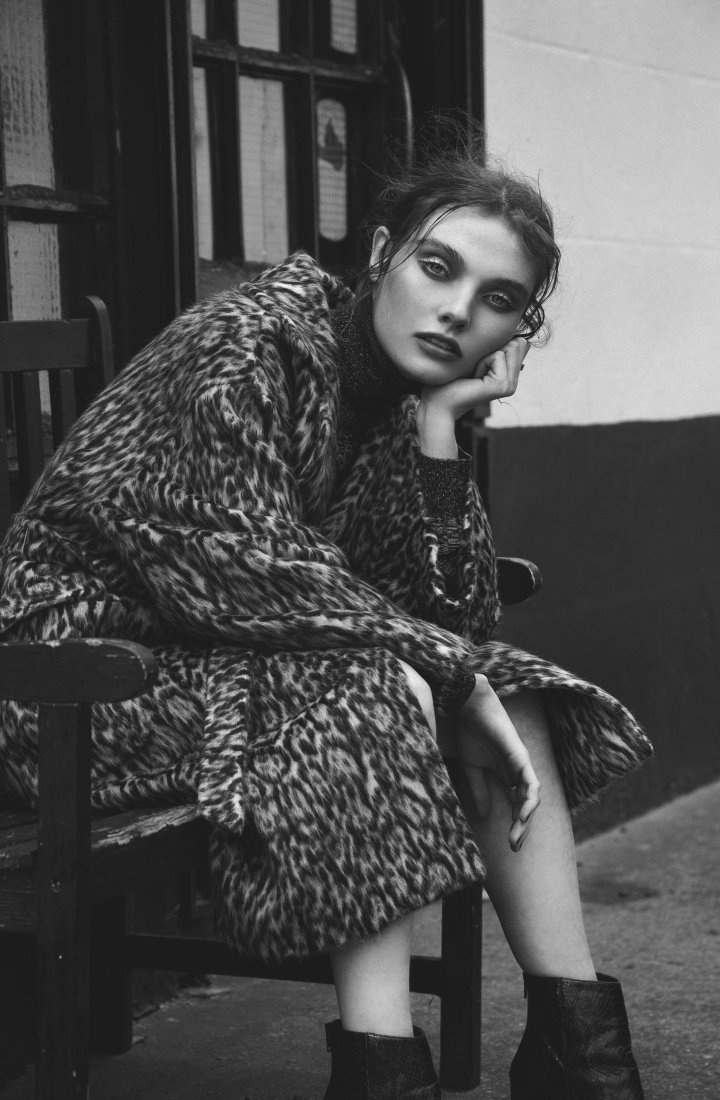 Black and white portrait of a woman sitting on a bench, wearing an animal print coat and boots in an outdoor setting with neutral tones.