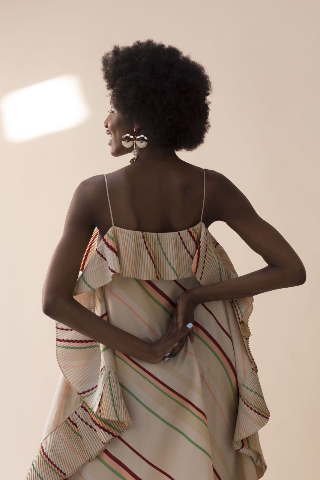 Back view of a Black woman with afro wearing a striped dress and large earrings against a beige background, lit by natural light.