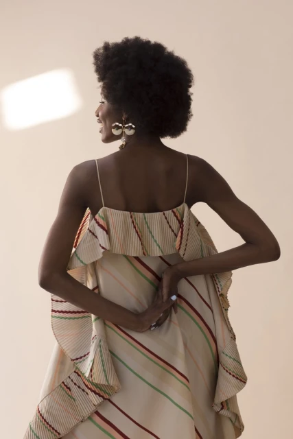 Back view of a Black woman with afro wearing a striped dress and large earrings against a beige background, lit by natural light.