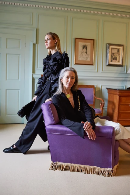 Two women pose indoors; one sits on a purple chaise lounge while the other stands in a black ruffled top and wide-leg pants.