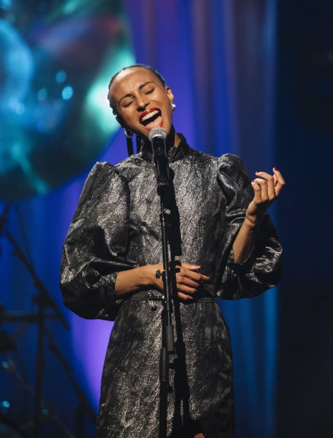 Elegant adult female singer with microphone on stage, wearing a dark dress in front of a blue and purple background.