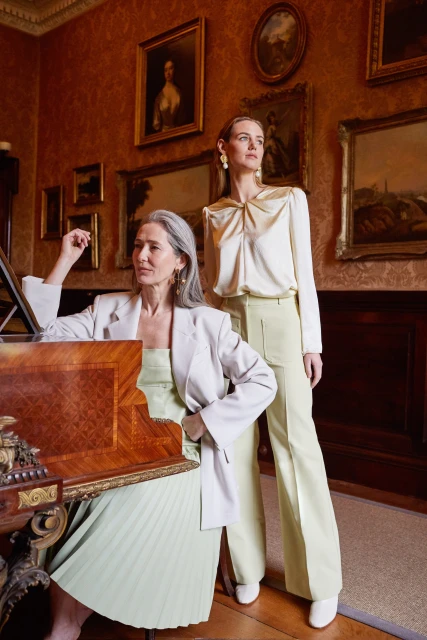 Two women in stylish attire pose in an ornate room with paintings. One is seated by a wooden desk, and the other is standing.