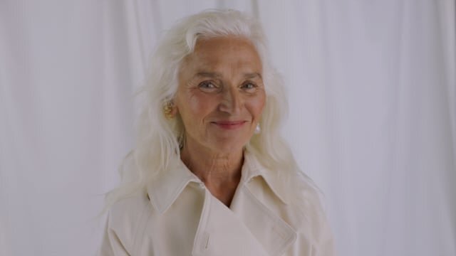 Elegant older woman with white hair smiles in a close-up portrait against a soft, draped white background, wearing a neutral trench coat.