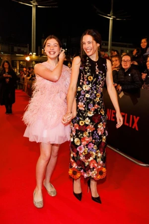 Two women on a red carpet at night. One woman wears a pink feathered dress, and the other wears a floral dress with black heels.
