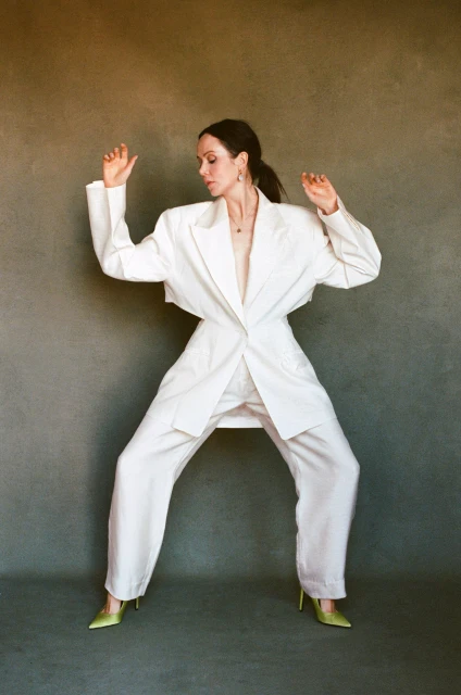 Woman in white suit and high heels strikes a dynamic pose against a neutral backdrop, showcasing a fashion-forward and confident style.