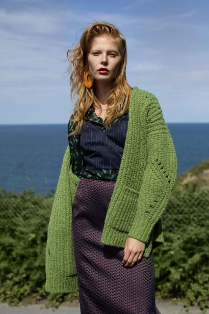 A fair-skinned woman with red hair and red lipstick stands in front of ocean wearing a green knit cardigan, patterned shirt, and skirt.