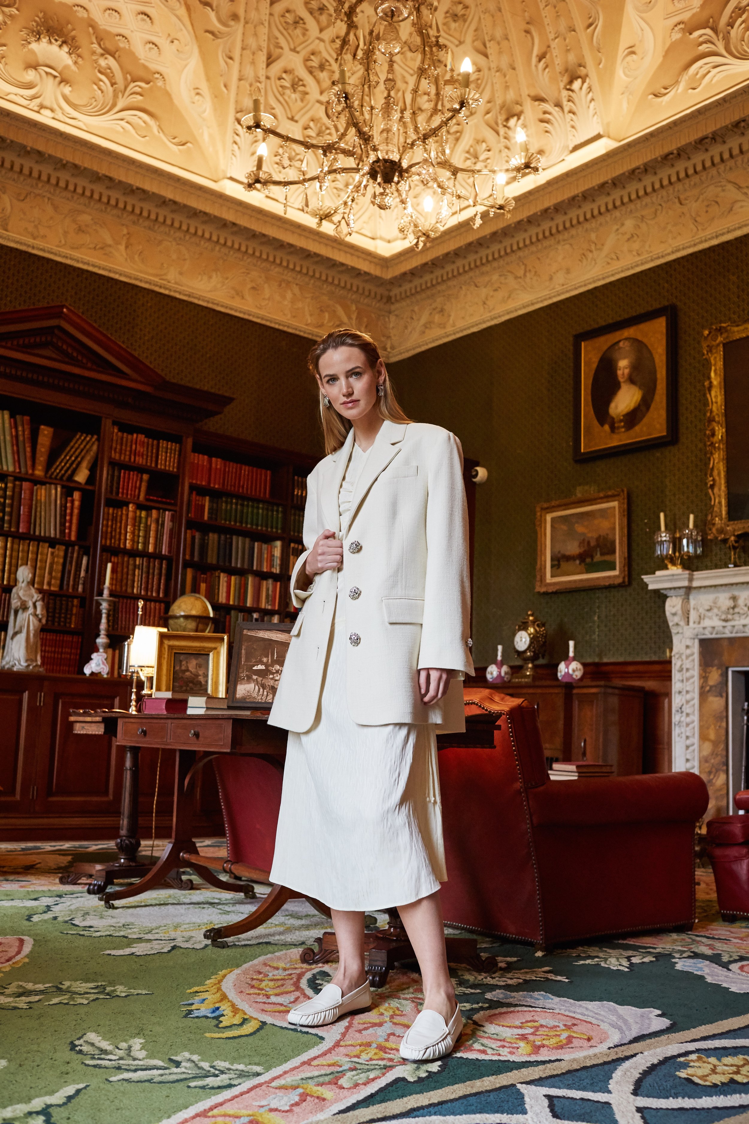 Indoor fashion shot of a woman in a cream-colored suit and skirt, posing in a library-style room with antique furniture and decor.