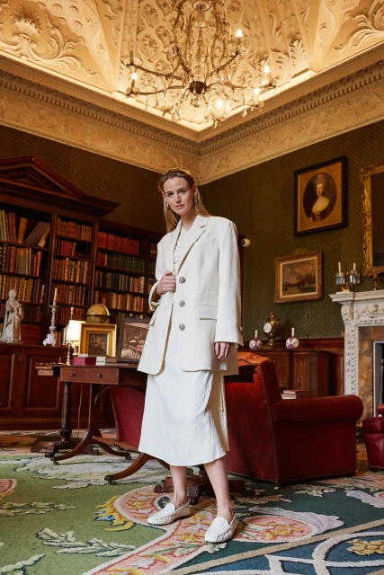 Indoor fashion shot of a woman in a cream-colored suit and skirt, posing in a library-style room with antique furniture and decor.