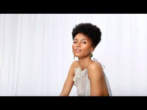 Portrait of a woman with short dark hair wearing a white ruffled top and green earrings against a white curtain background.