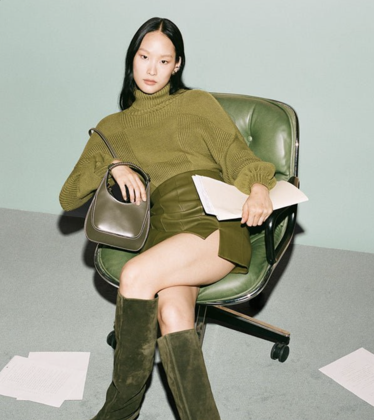 A stylish Asian woman sits in an olive green office chair wearing matching separates with a handbag and knee-high boots. 