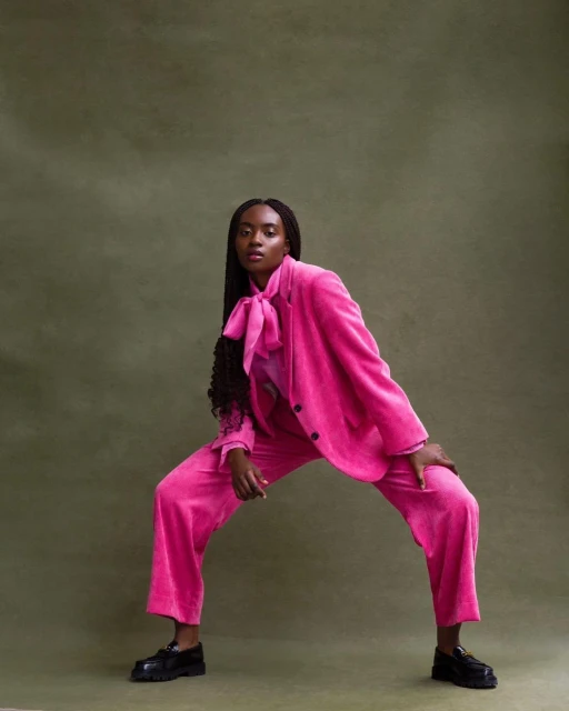 Fashionable woman in pink velvet suit and black loafers poses on an olive green background in a studio fashion shot.
