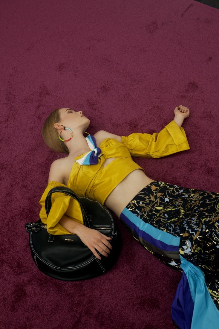 Model in yellow top and patterned pants poses with a black handbag on a maroon carpet, showcasing a high-fashion, editorial style.