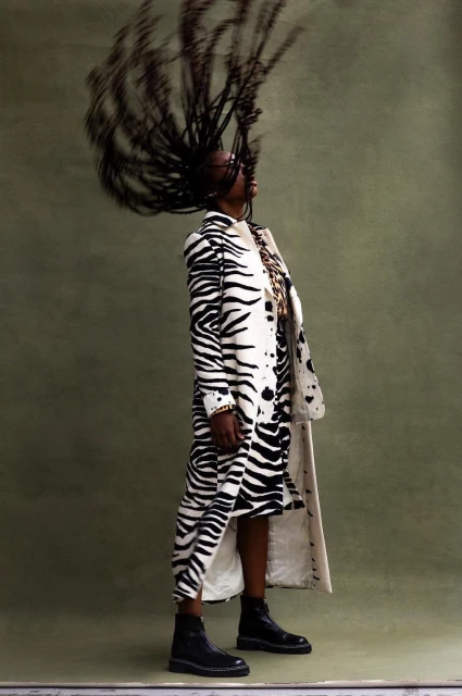 A woman wearing a white and black zebra-print coat and dress stands in a photo studio while whipping her long hair.