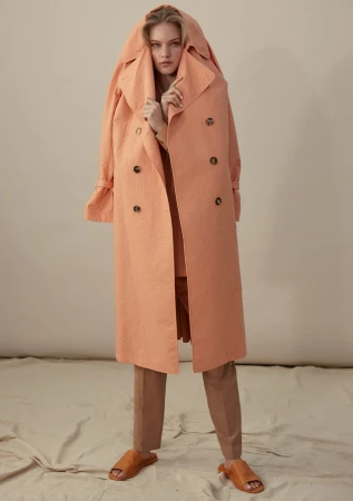 Model in peach trench coat with hood pulled over head and brown slides against a plain cream backdrop