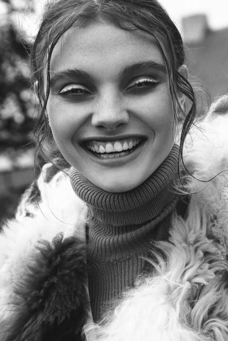 Close-up portrait in black and white of a young woman smiling and wearing a fur coat and turtleneck sweater outdoors.