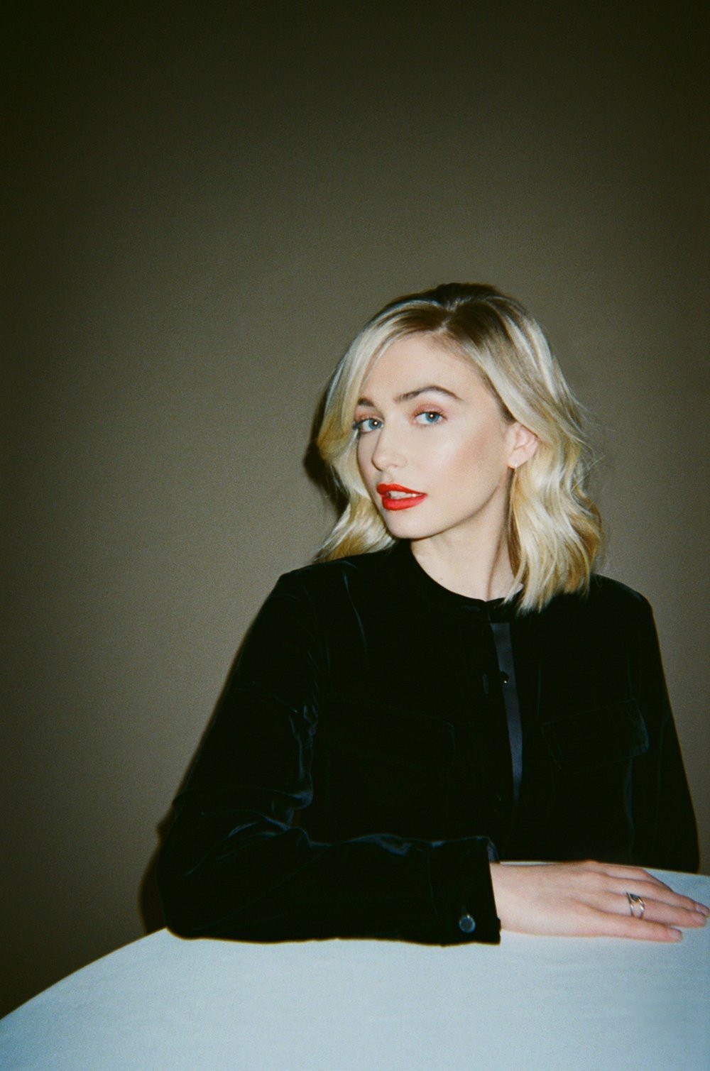 Blonde woman with red lipstick poses indoors in a black velvet jacket on white table. Fashion, beauty, portrait, and lifestyle.