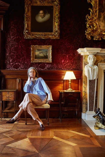 Adult woman in a blue shirt seated in an ornate room with wood paneling, paintings, parquet floors, and antique furniture.